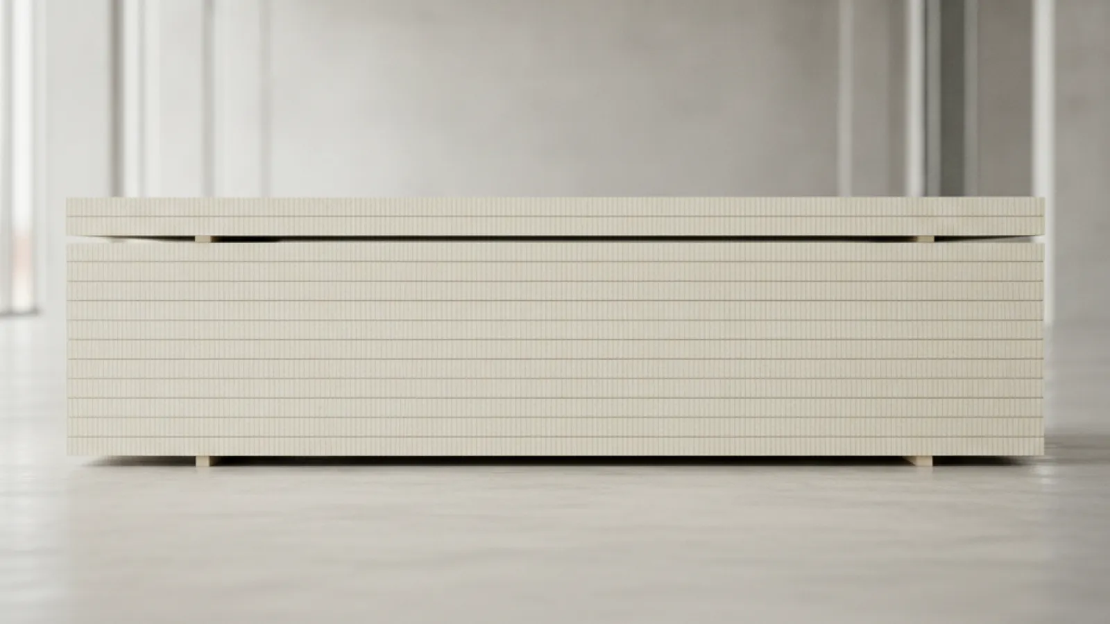 A low, long unit made of stacked light beige panels with a fine vertical texture and small wooden feet is centered on a light gray concrete floor in a brightly lit, empty room with large windows and concrete walls.